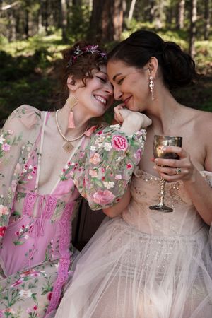 Two people in elegant dresses share an intimate moment while enjoying wine at an outdoor celebration.