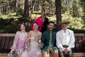 Wedding party members in medieval-inspired attire pose on a wooden bench in a forest setting.