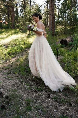 A bride in a flowing off-shoulder wedding gown holds a bouquet while standing in a sunlit forest setting.