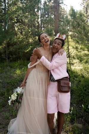 Two people share a joyful embrace in a forest setting, one wearing a flowing wedding dress and the other in pink casual attire.