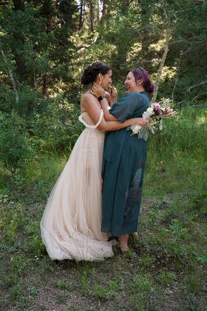 Two people embrace in a romantic woodland setting, one wearing a flowing blush gown and one in teal dress holding flowers.