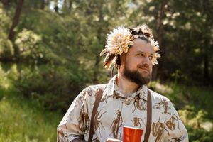 A person in a floral crown and patterned shirt gazes into the distance in a sunlit forest.