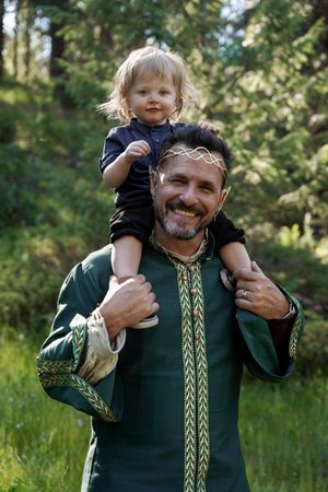 Adult wearing decorative green medieval-style tunic carries small child on shoulders in outdoor forest setting.