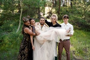Group lifting and carrying someone in a wedding dress through a wooded outdoor setting.