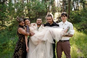 Wedding party members lift and carry someone in a white dress outdoors.