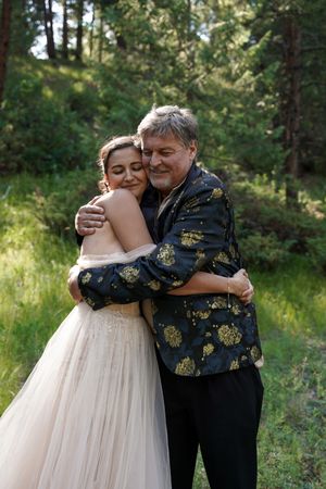 A tender embrace between two people in formal wear against a forest backdrop at an outdoor celebration.