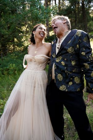 Couple shares joyful laughter in a forest setting with bride in flowing gown and groom in patterned jacket.