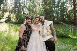 Wedding party poses together in a sun-dappled forest clearing, with bride in flowing white gown.