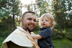 A playful moment between an adult and child in medieval-inspired costume against a forest backdrop.