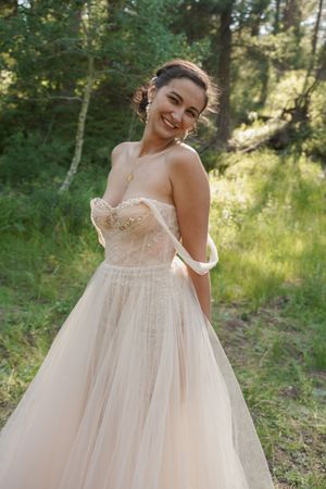 A flowing blush pink wedding dress with delicate lace details is showcased in a sun-dappled forest setting.