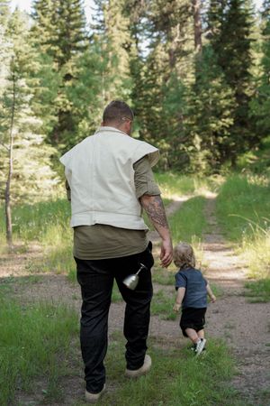 Adult and child holding hands while walking together on a forest trail surrounded by evergreen trees.