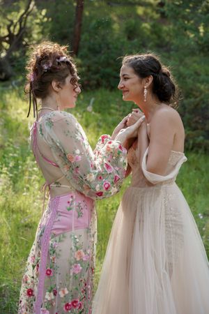 Two people in elegant dresses share an intimate moment in a sunny garden setting.