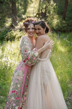 Two people embrace in a forest setting, one wearing a floral dress and one in a tulle wedding gown.
