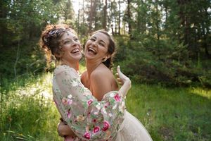 Two people in wedding attire share a joyful embrace outdoors in a sunlit wooded setting.