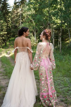 Two people in flowing dresses walk through a sunlit forest path together.