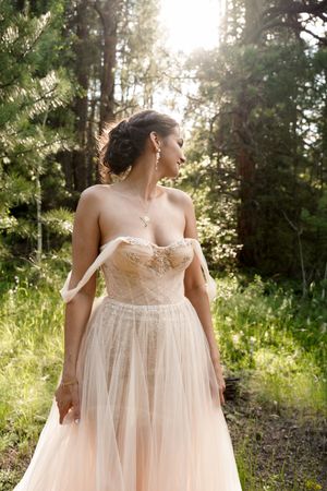 A bride in a flowing blush tulle gown poses in a sunlit forest setting.