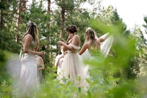 Three women in white dresses gather in a sunlit forest clearing during a wedding celebration.