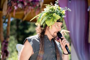 A performer in medieval attire wearing a floral crown speaks into a microphone during an outdoor celebration.