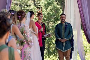 An outdoor wedding ceremony takes place under white and purple draped fabric in a lush garden setting.