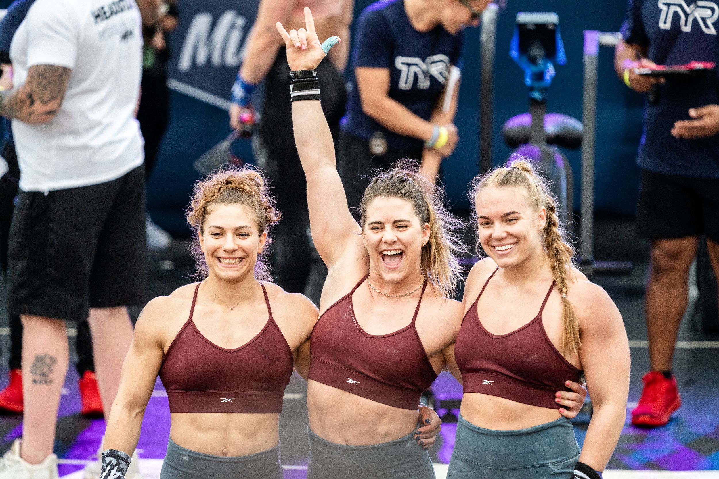 Three athletes in matching burgundy sports bras celebrate with raised arms during a fitness competition.