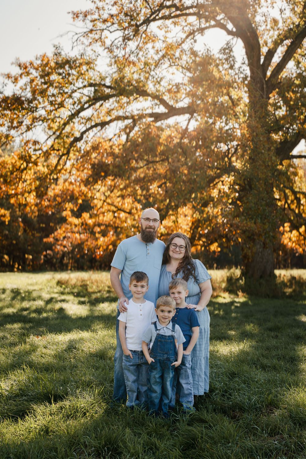 A family sits together in a grassy field during golden hour as autumn sunlight filters through trees behind them.