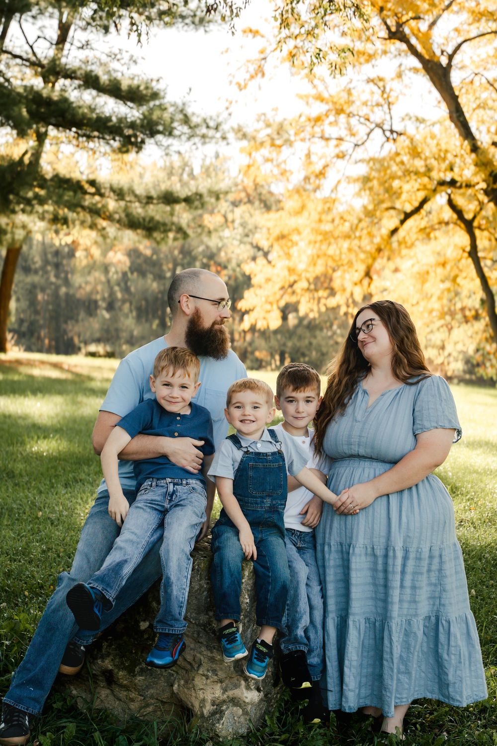 Family wearing coordinated denim and blue outfits poses together in a sunlit autumn park setting.