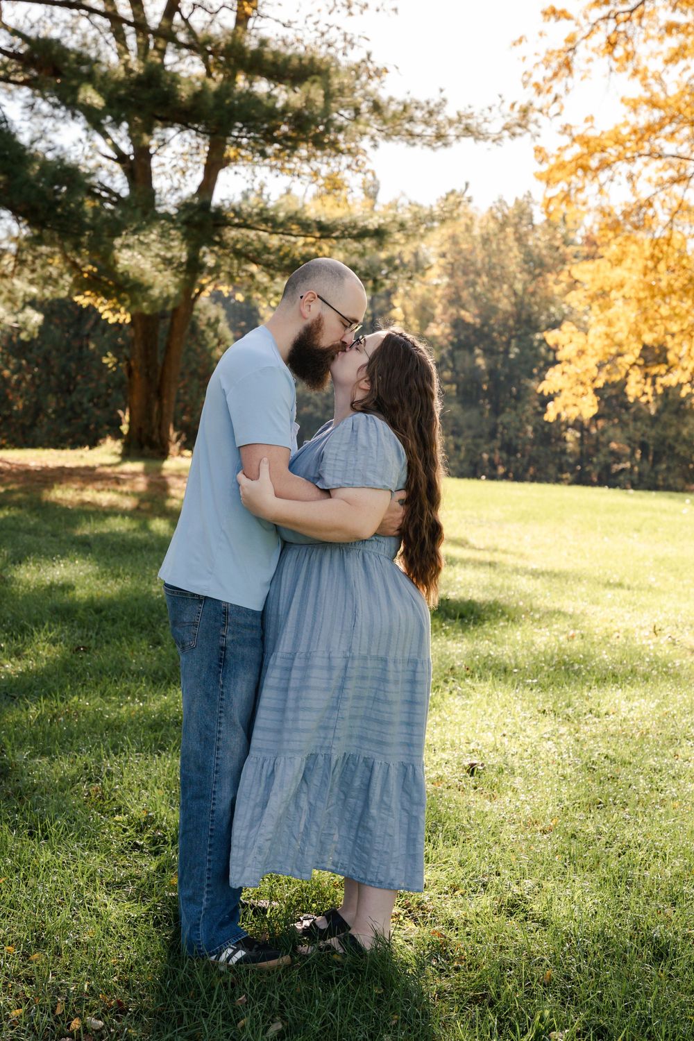 A romantic couple embraces in a sunlit meadow during golden hour while wearing casual blue and white outfits.
