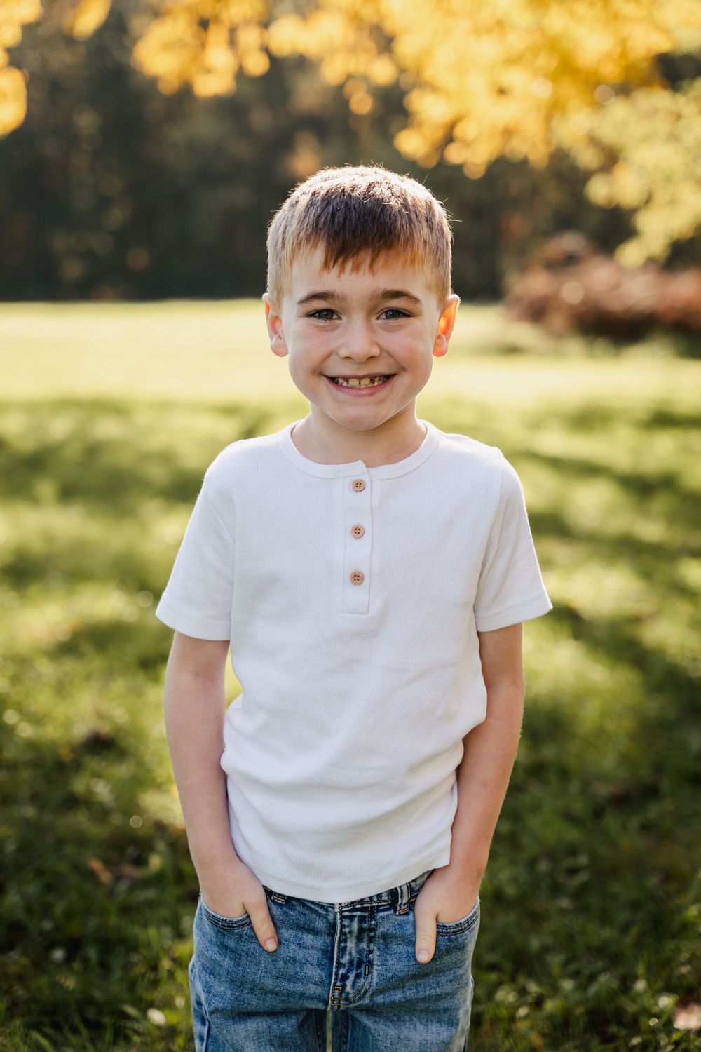 A young child wearing a white henley shirt and blue jeans stands outdoors in golden sunlight.