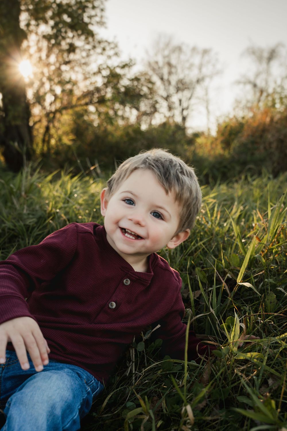 A child in a burgundy sweater sits in tall grass during golden hour as sunlight filters through autumn trees in the background.