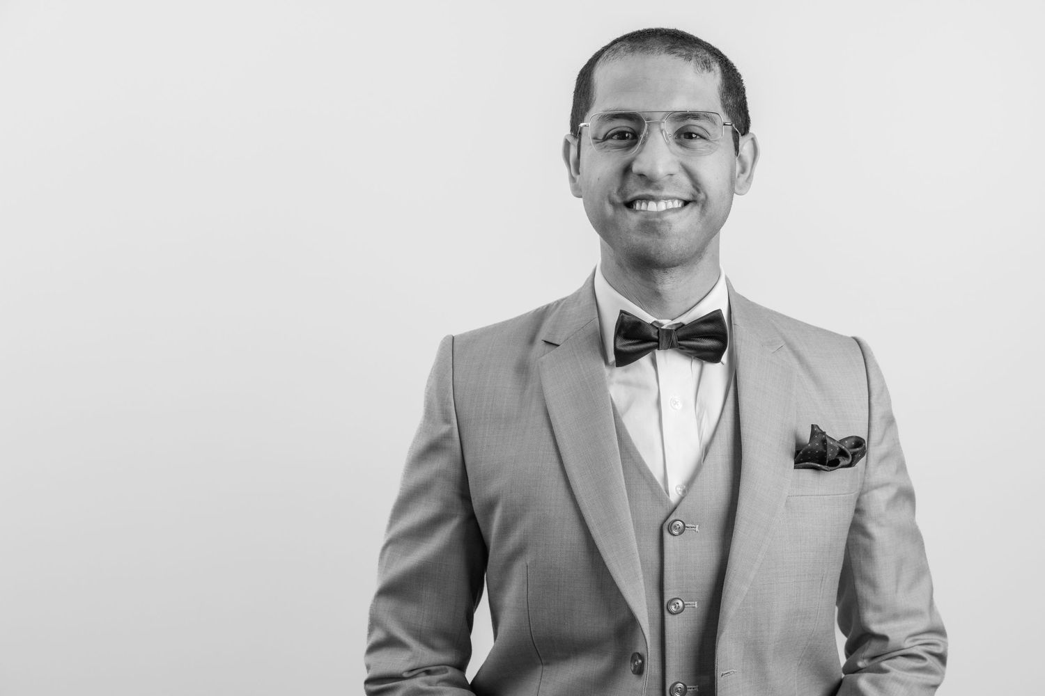 A smiling person in a light gray three-piece suit with bowtie and pocket square poses against white background in black and white.