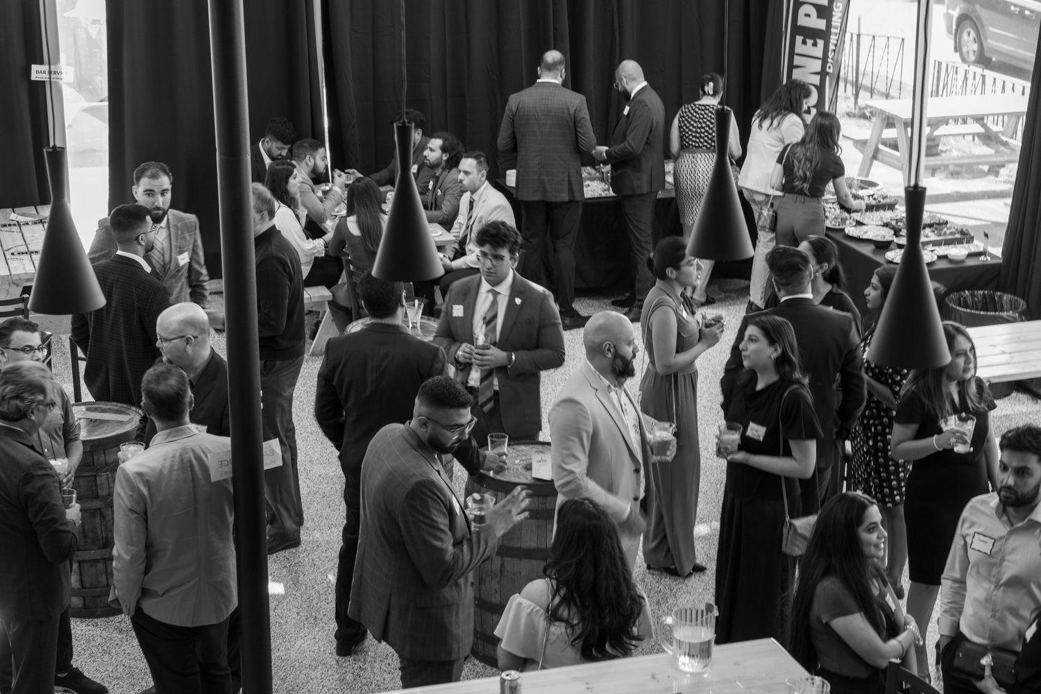 A black and white photo of guests socializing at a formal event with wine bottles on display.