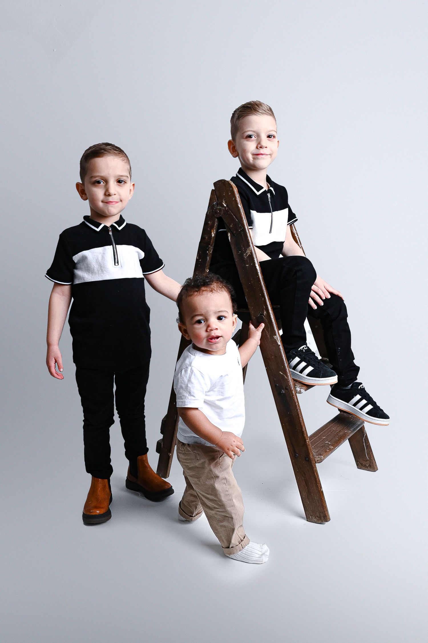 Series of studio photos showing toddler in black outfit posing with wooden ladder against white backdrop.