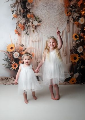 Children in white dresses pose playfully against a backdrop with orange sunflowers and macrame details.