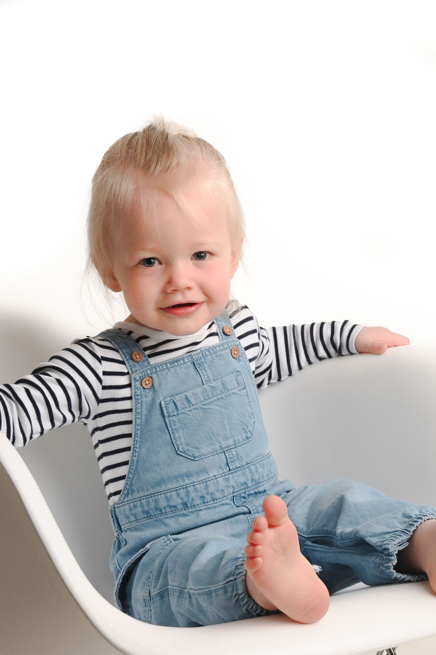 Happy baby in striped shirt and denim overalls sitting in white chair.
