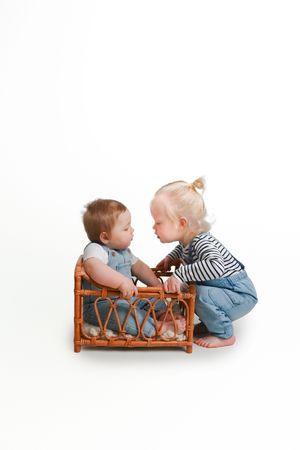 Two babies in denim overalls interacting with wicker furniture against white background.