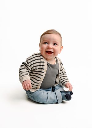 Baby wearing striped sweater and jeans sitting and smiling on white background.