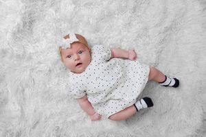 Baby in white spotted outfit with black shoes lying on white rug.