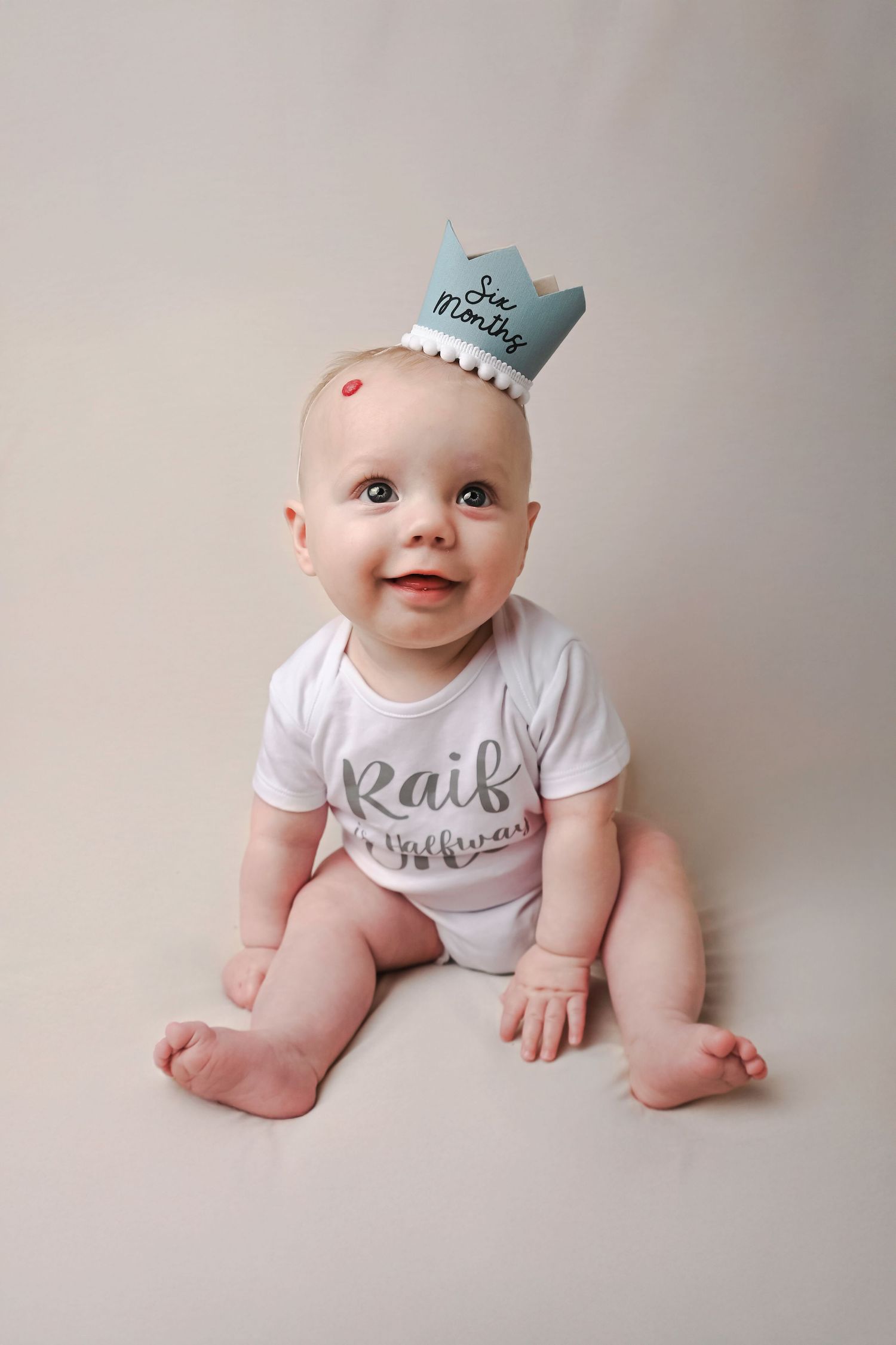 Baby in white onesie with blue crown accessory posing on gray background.