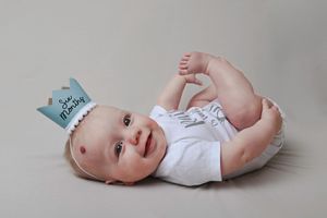 Baby wearing white onesie lying on gray background with blue crown accessory.
