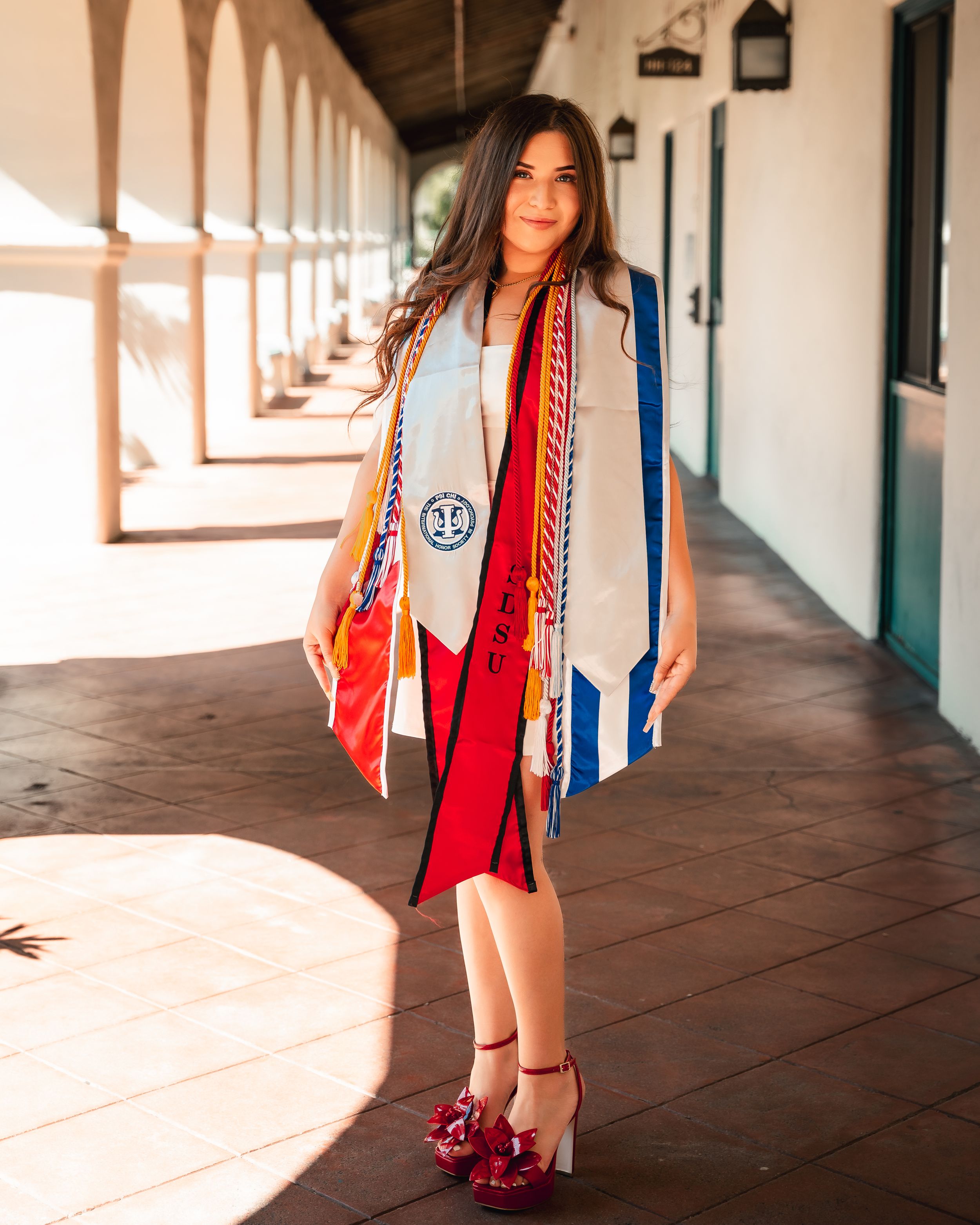 Graduate in white dress and colorful stoles poses in Spanish colonial style hallway.