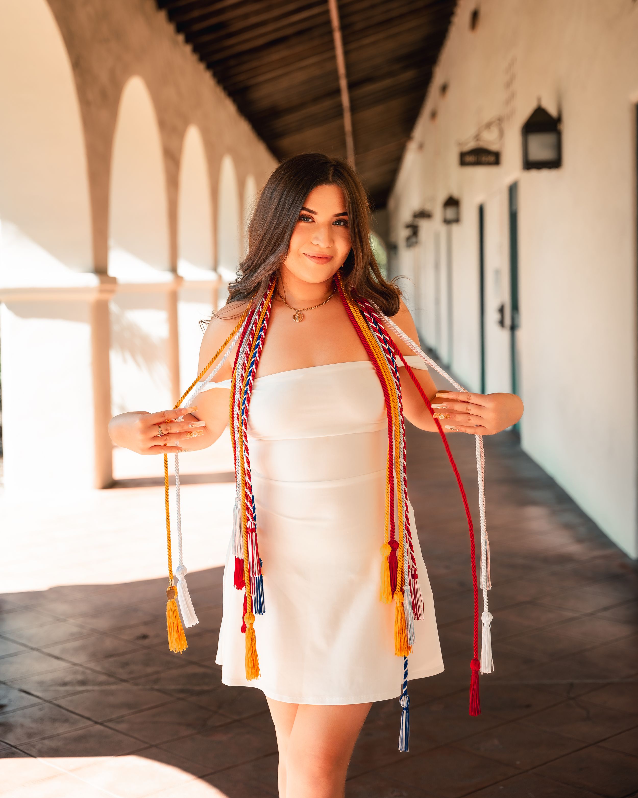 Graduate wearing white dress and graduation stoles stands in sunlit corridor.