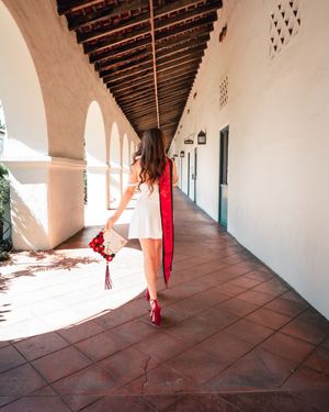 Graduate in white dress and red stole dances through sunlit Spanish colonial corridor.