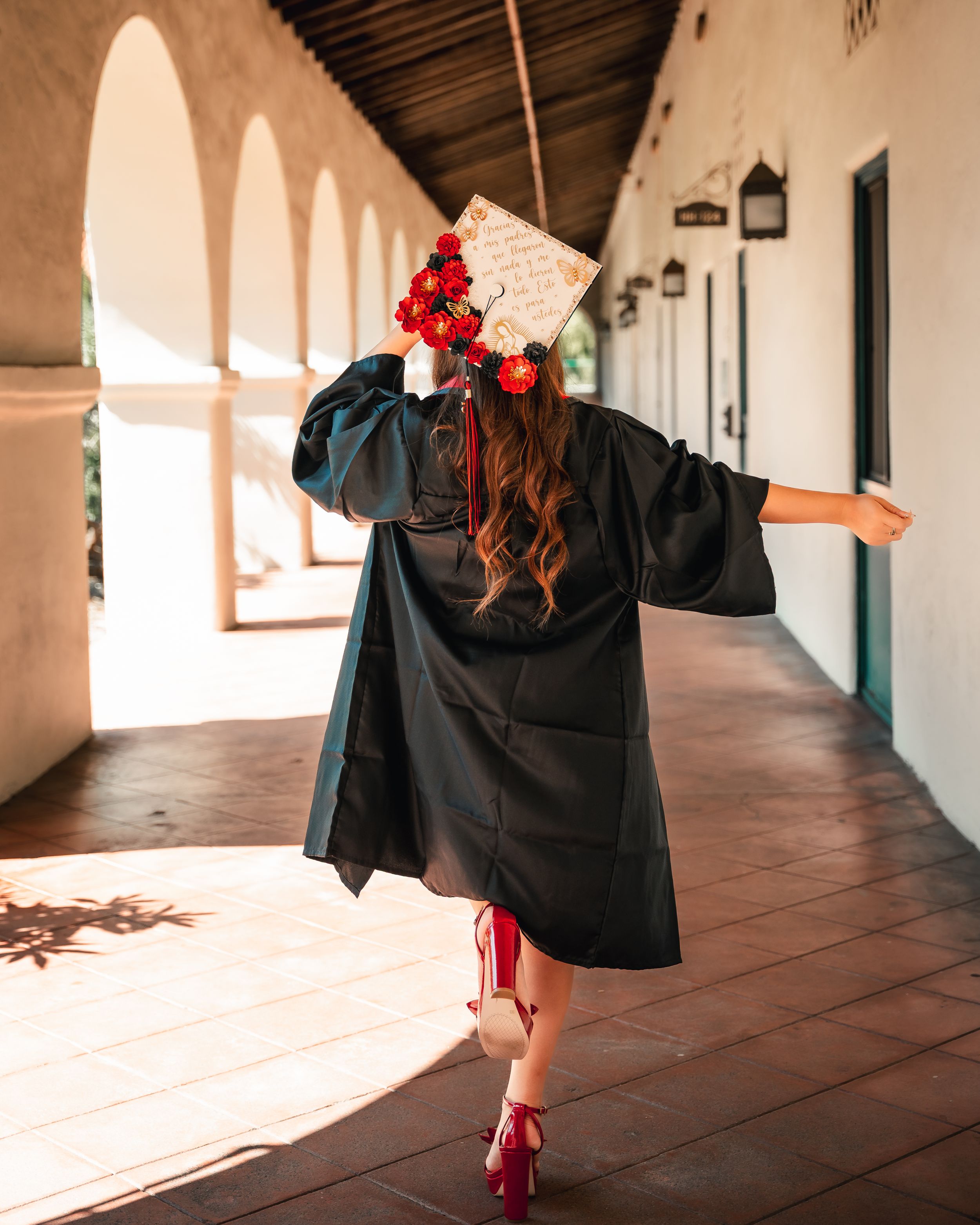Graduate in black cap and gown twirls in sunlit hallway with decorated cap.