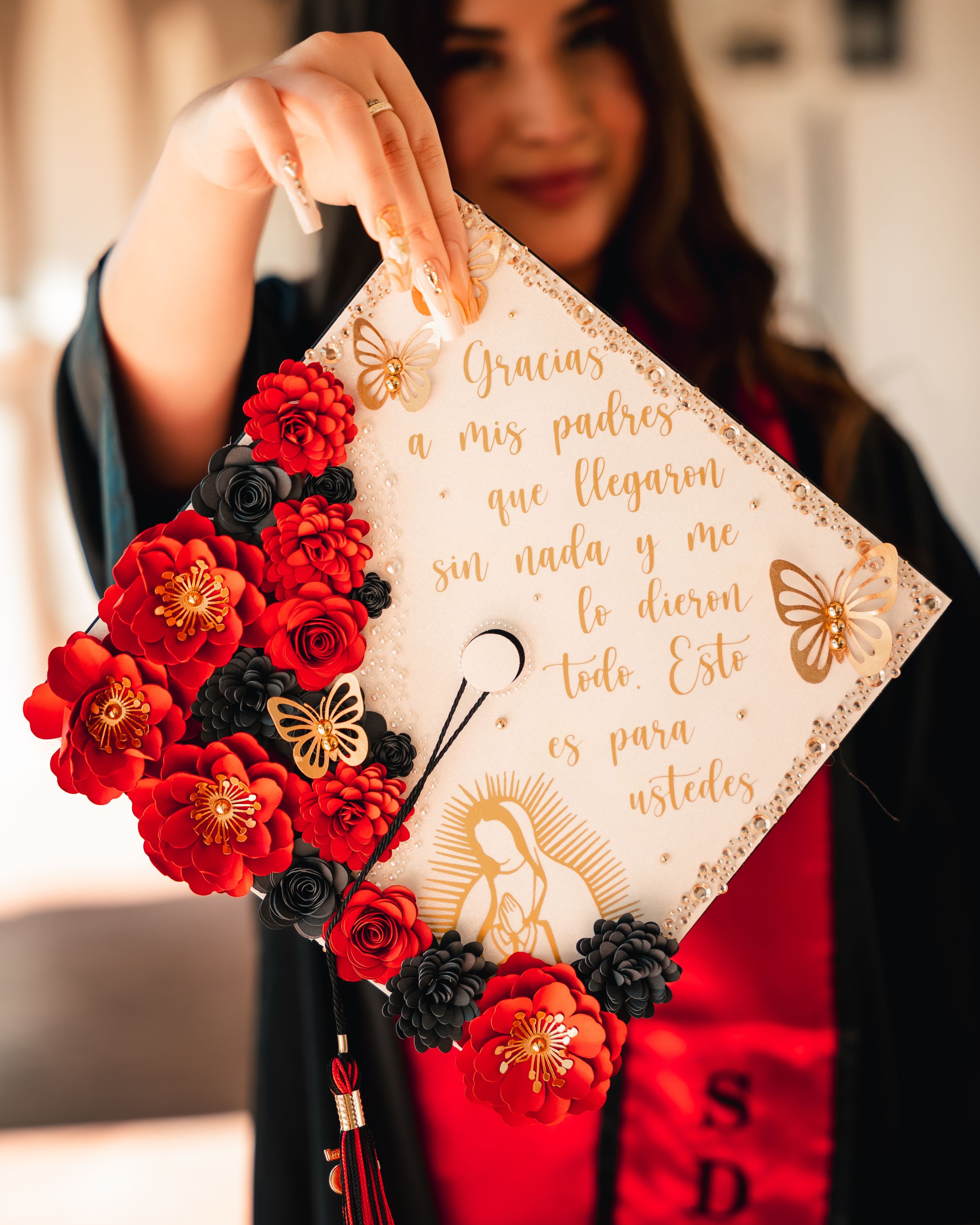 Decorated graduation cap with red flowers and Spanish text against architectural background.