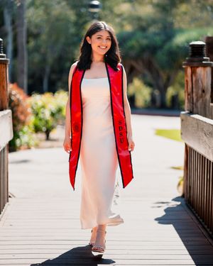 Graduate in white dress and red stole poses on wooden bridge outdoors.