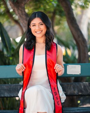 Graduate in white dress and red stole celebrates on outdoor bench.