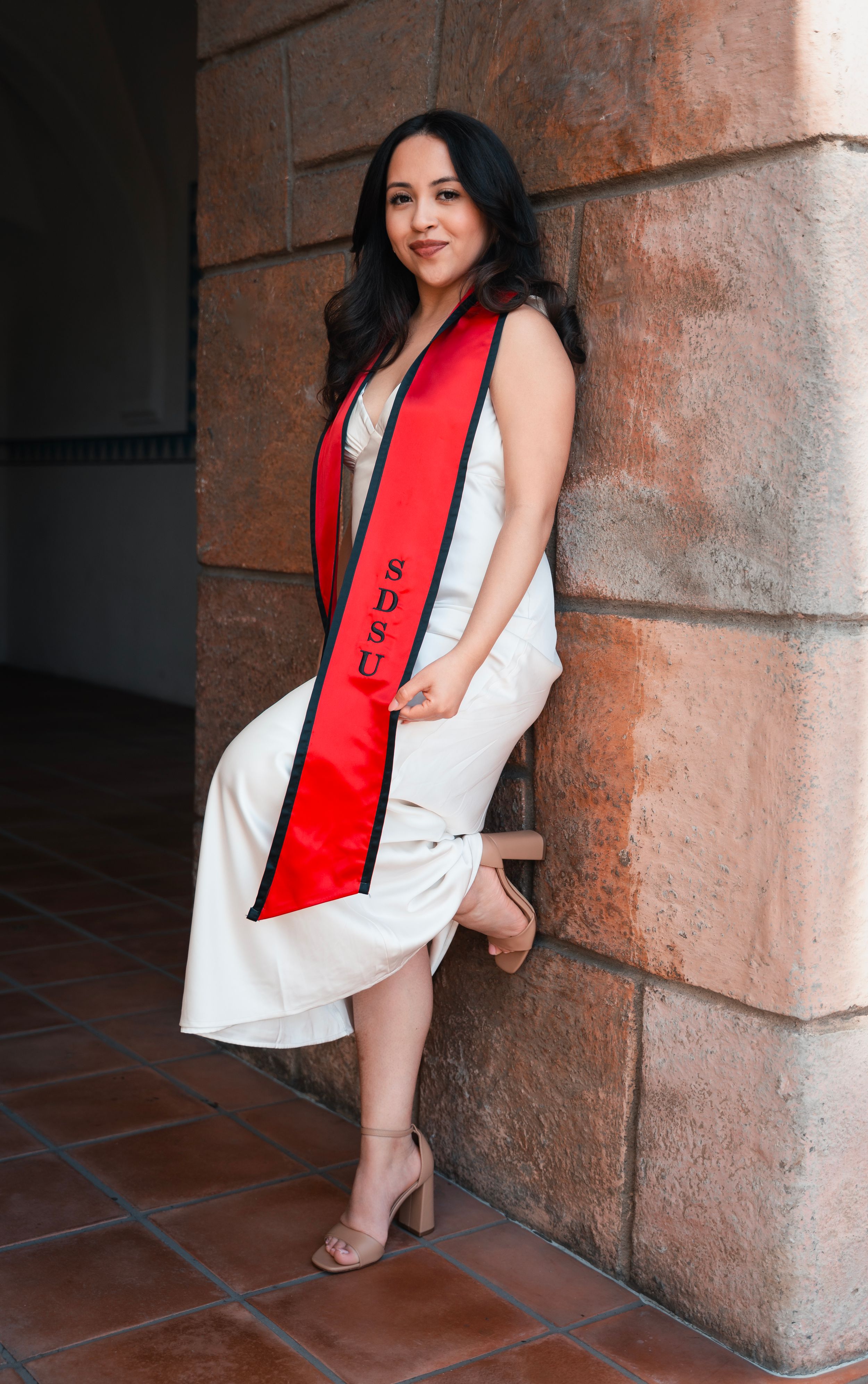 Graduate posing in white dress and red stole against stone pillars in an outdoor setting.