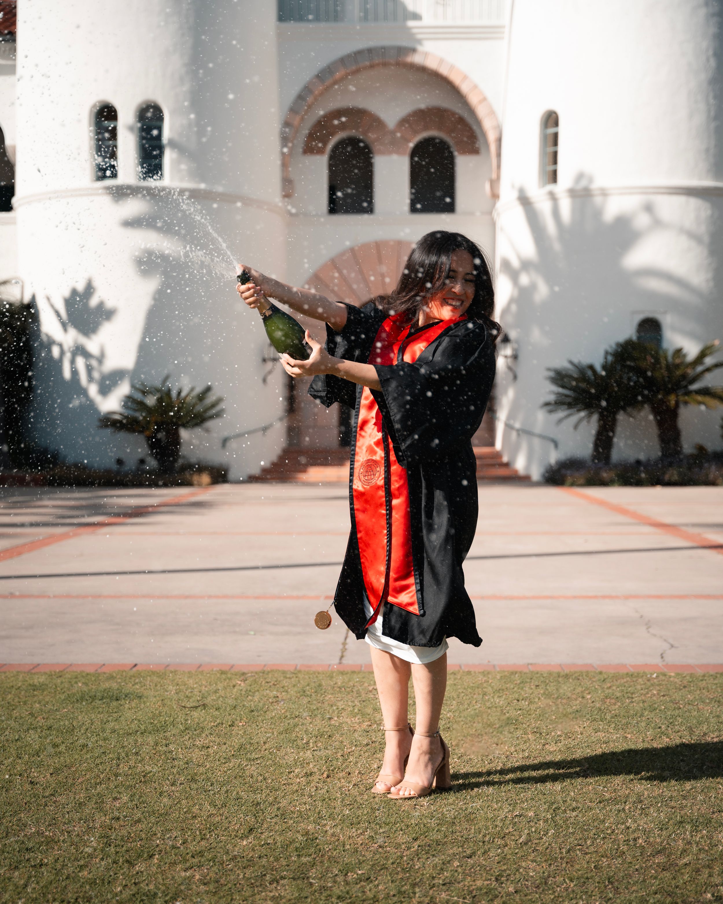 Celebratory graduation poses on grass with white building and palm tree shadows in background.
