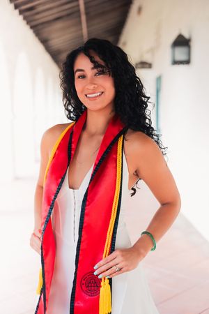 Smiling graduate wearing red and gold stole poses against white wall.