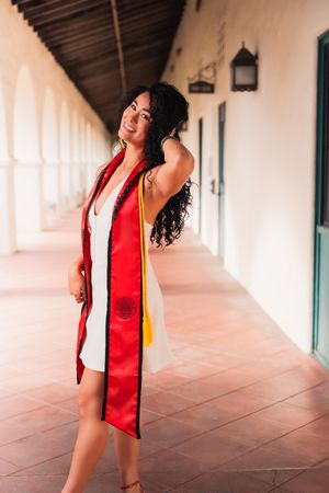 Graduate in white dress and red graduation stole poses joyfully in outdoor hallway.